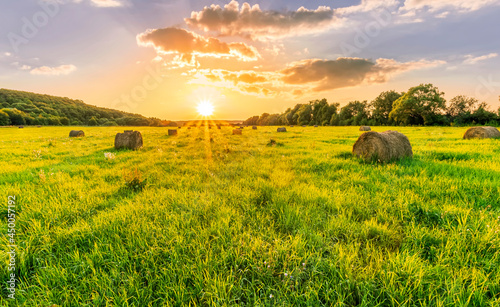 Scenic view at sunset or sunrise in green shiny field with hay stacks  bright cloudy sky  golden sun rays  summer valley landscape