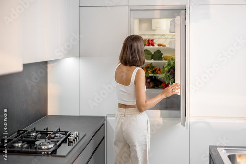 Young woman standing near the fridge full of fresh vegetables at modern kitchen. Healthy vegan eating concept photo