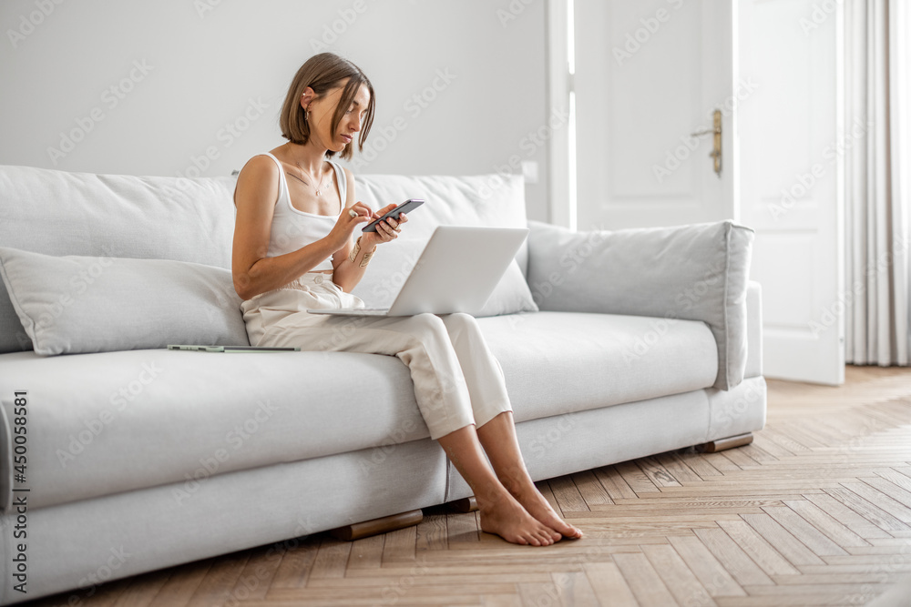 Young woman works with laptop and phone while sitting on the comfortable couch at home. Work from home concept Young woman works with laptop and phone while sitting on the comfortable couch at home. Work from home concept