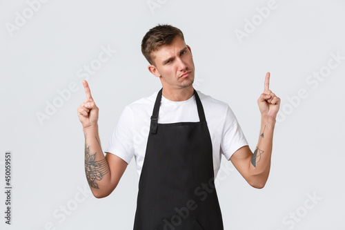 Small business owners, coffee shop and staff concept. Skeptical and dispelased barista in black apron judging, looking disapponted and annoyed, pointing fingers up and scolding someone photo