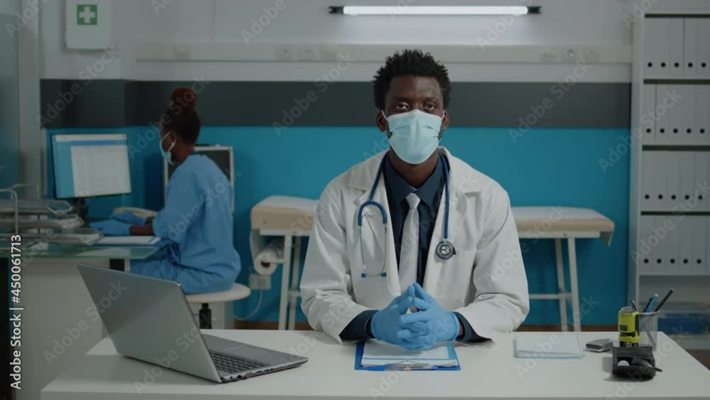 Portrait of african american doctor looking at camera wearing white coat in office at medical facility. Black medic sitting at desk with laptop, tools, stethoscope, and documents Portrait of african american doctor looking at camera wearing white coat in office at medical facility. Black medic sitting at desk with laptop, tools, stethoscope, and documents