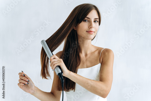 Waist up portrait of the contented young woman using a hair straightener in the bathroom while looking at the camera with pleasure smile. Woman appearance concept