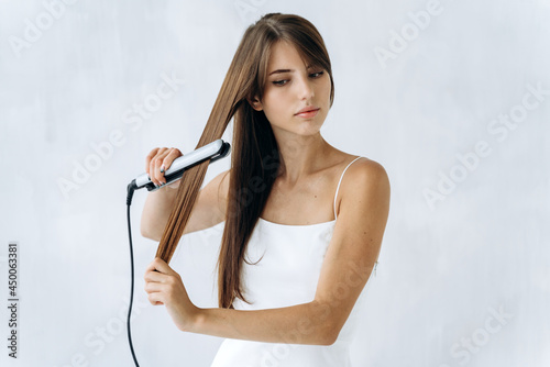 Adorable young woman straightening her hair with a flat iron and looking aside with calm emotions. White wall at the background. Woman hair concept photo