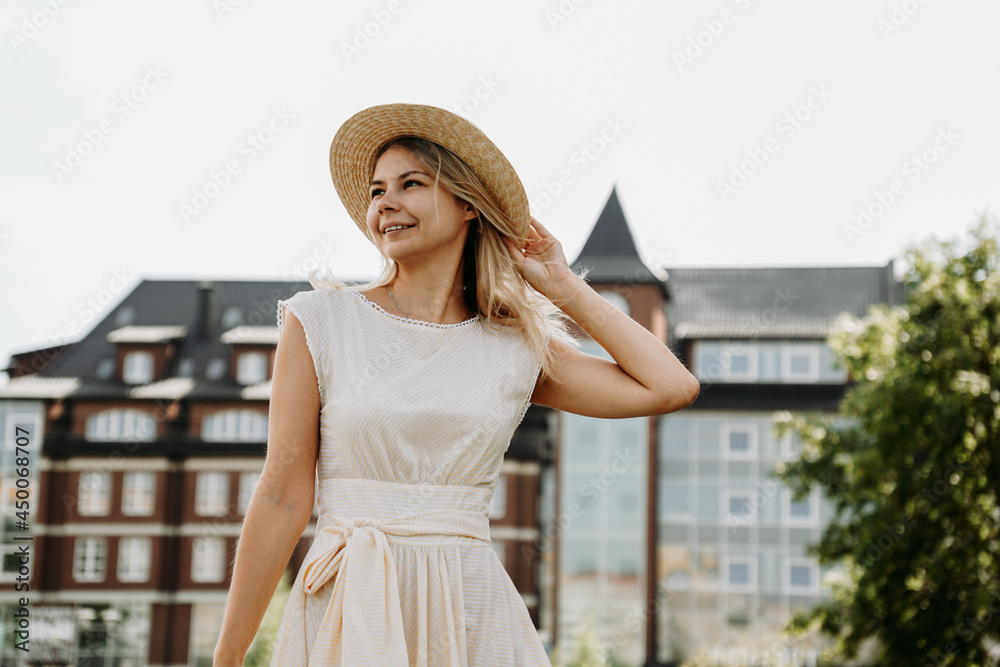 A beautiful blonde walks through a European city. Woman in white dress and straw hat, she smiles