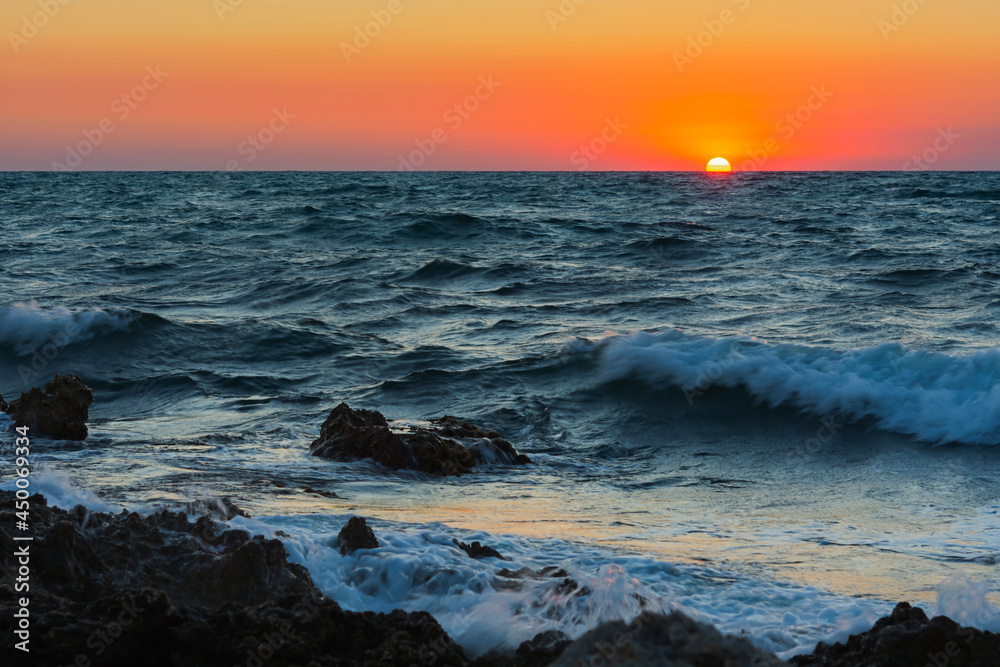 Sunset and storm at sea. Big waves against the setting sun. A summer storm in the ocean. Beautiful sea spray with foam breaks on the rocky shore. Beautiful natural background. The orange setting sun. Sunset and storm at sea. Big waves against the setting sun. A summer storm in the ocean. Beautiful sea spray with foam breaks on the rocky shore. Beautiful natural background. The orange setting sun.