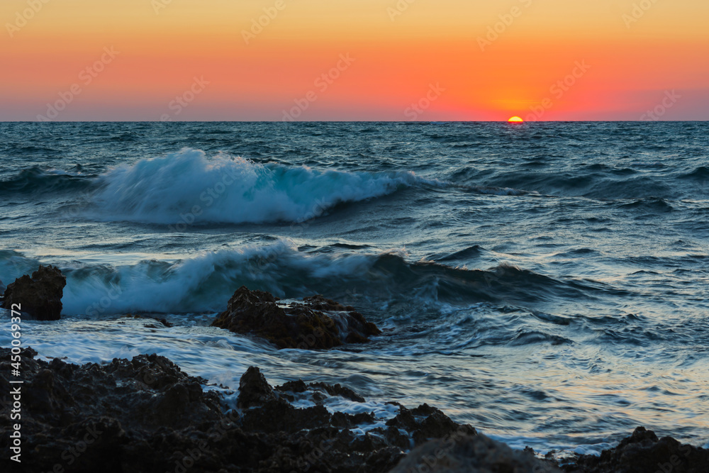 Sunset and storm at sea. Big waves against the setting sun. A summer storm in the ocean. Beautiful sea spray with foam breaks on the rocky shore. Beautiful natural background. The orange setting sun.