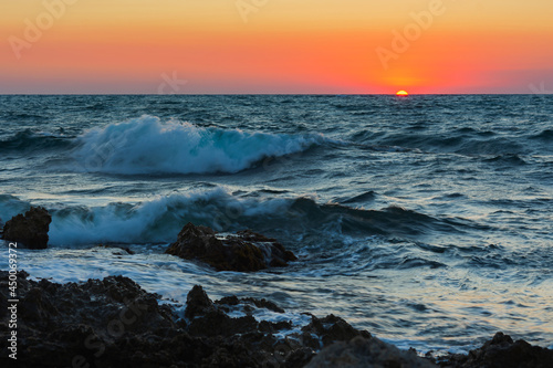 Sunset and storm at sea. Big waves against the setting sun. A summer storm in the ocean. Beautiful sea spray with foam breaks on the rocky shore. Beautiful natural background. The orange setting sun.