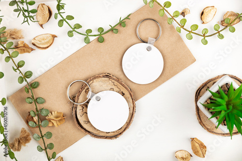 From above modern keychain mock ups with blank round pendants placed on piece of wood and rectangular cardboard near green plants and dried leaves on white background. Key chain mock up photo