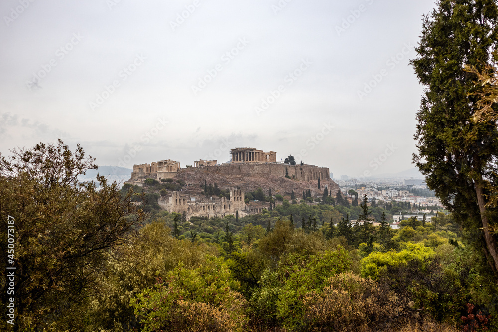 Acropolis hill (Parthenon, Propylaea, Temples, Odeon of Herodes Atticus) view through summer greenery. Athens ancient historical landmark in city center from Filopappou Hill on cloudy day