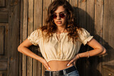 Summer portrait of a curly pretty woman with fashion sunglasses in fashionable clothes stands and looks at the camera near a wooden wall on the beach at sunshine