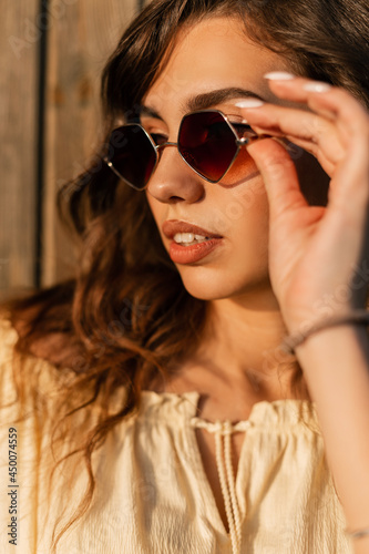 Fashion summer portrait of stylish pretty woman with curly hair put on a sunglasses near wooden wall at sunset