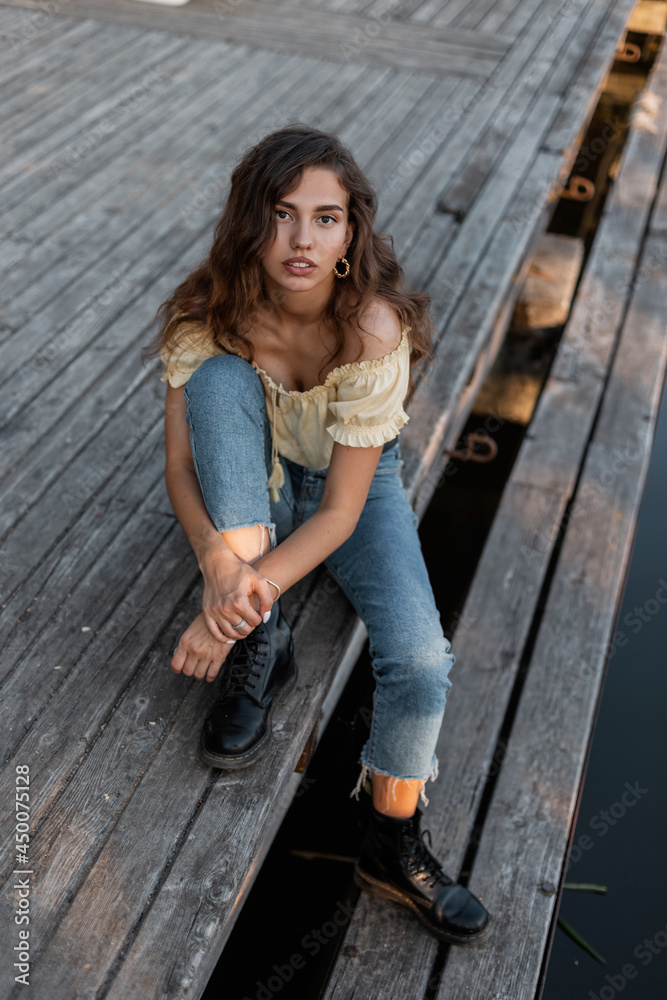 Beautiful curly woman in fashion denim clothes with jeans, blouse and shoes sits on a wooden pier near the lake. Beautiful curly woman in fashion denim clothes with jeans, blouse and shoes sits on a wooden pier near the lake.