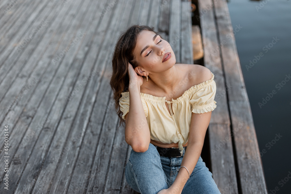 Sensual beautiful young woman in trendy summer clothes with jeans and a yellow top is resting and sitting on a wooden pier near the sea