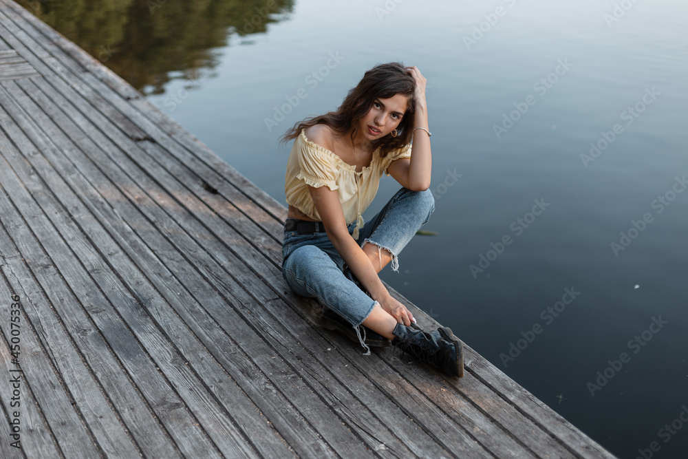 Fashionable beauty woman with curly hair in blue jeans with yellow top blouse and boots sits and rest on a vintage wooden pier near lake