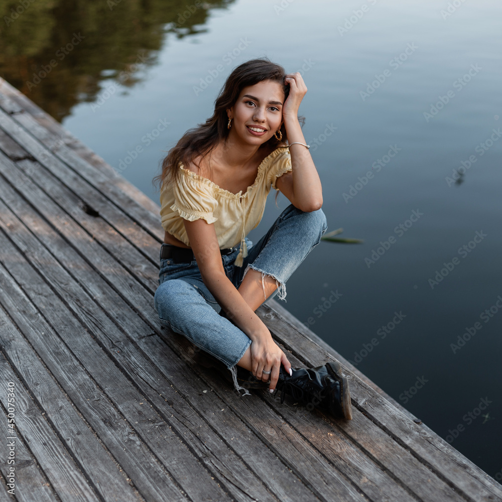 Pretty young happy curly woman with a smile in stylish denim clothes sits and rest on the wooden pier by the lake. Summer vacations