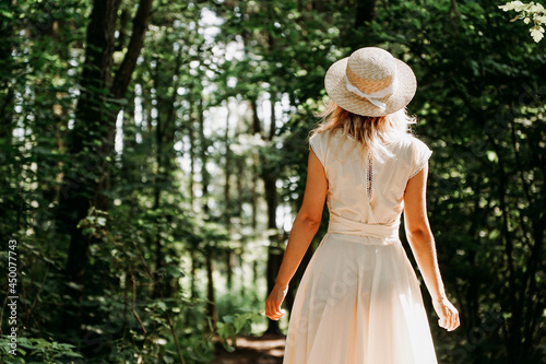 Beautiful young woman in a straw hat and white dress in a green park or forest on a summer day. Back view of a young happy woman