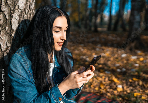 Portrait of a happy young woman reading a text message on a mobile phone by a tree in an autumn park. Chatting with friends using your smartphone.