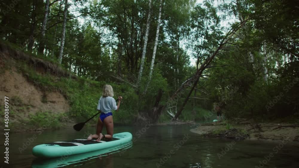 Young woman paddling on surfing board, standing on knees, man swimming after rowing on paddle board, back view. Female coach rows on surfboard. Sports lesson for beginners on water. Young woman paddling on surfing board, standing on knees, man swimming after rowing on paddle board, back view. Female coach rows on surfboard. Sports lesson for beginners on water.