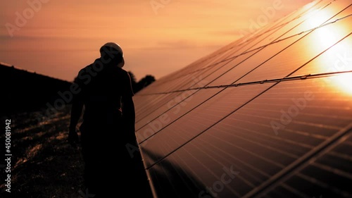 Assistance technical worker in uniform is checking an operation and efficiency performance of photovoltaic solar panels. Unidentified solar power engineer touches solar panels with his hand at sunset photo
