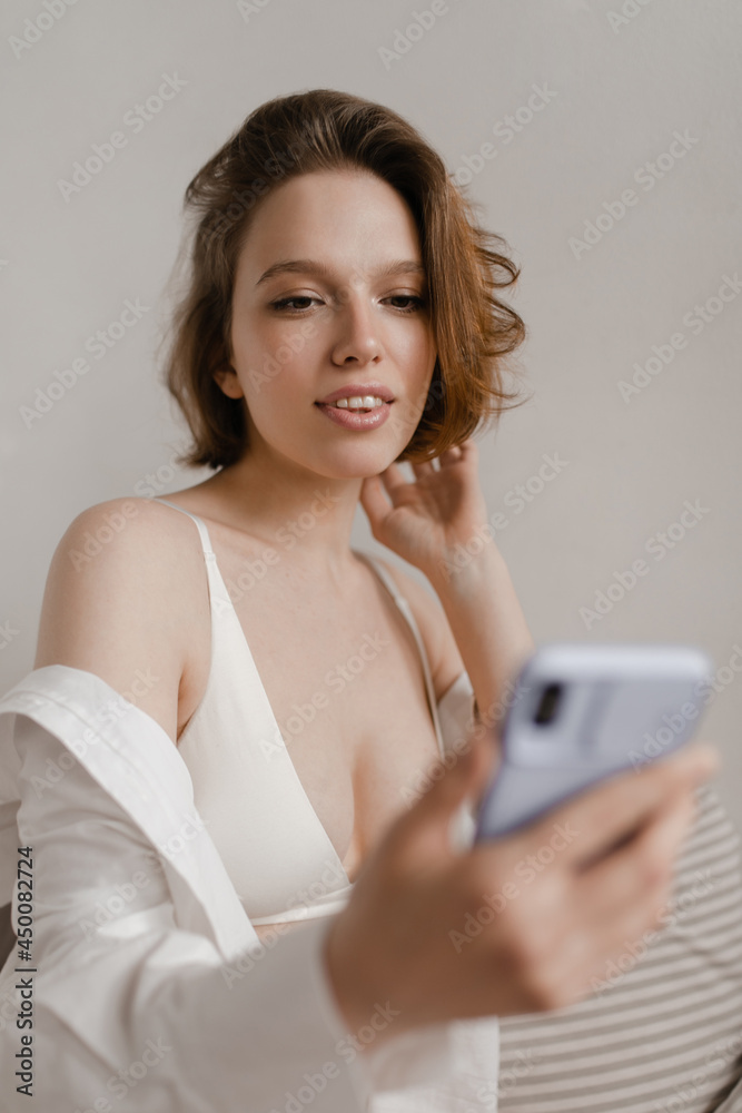 Young lady looking at phone in hands. Gorgeous dark-haired girl, wearing white bra and shirt, holding mobile and looking down isolated over light background indoors