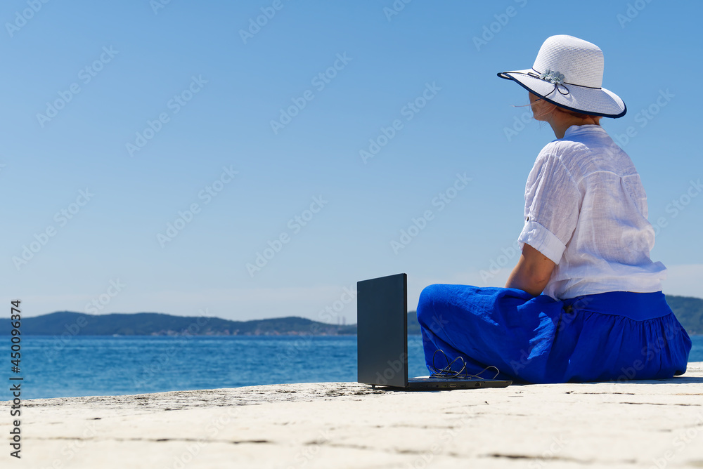 business woman freelancer sits on a pier by the sea and works on a laptop. dream job, freelance paradise, remote work outside the office