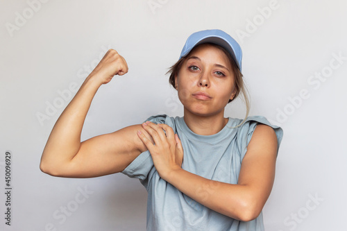 Tela Strong confident caucasian young blonde woman in a gray t-shirt and cap raises arm and shows bicep isolated on a white background