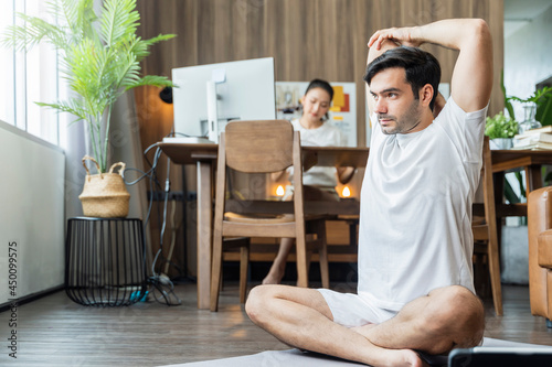 young asian male husband train yoga exercise workout praticing feomr online lesson at living room with wife working at behind,working from home stay home state order concept