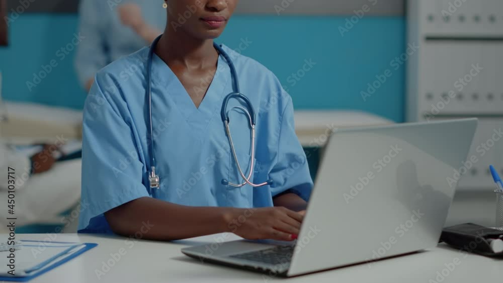 Close up of african american nurse using laptop computer on white desk at healthcare clinic. Black medical assistant typing on device while looking at camera with stethoscope and uniform