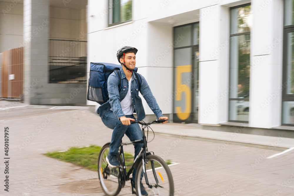 Joyful handsome young delivery man with thermo backpack riding bicycle in city street on blurred background of office building. Courier male wearing protective helmet delivery food to client