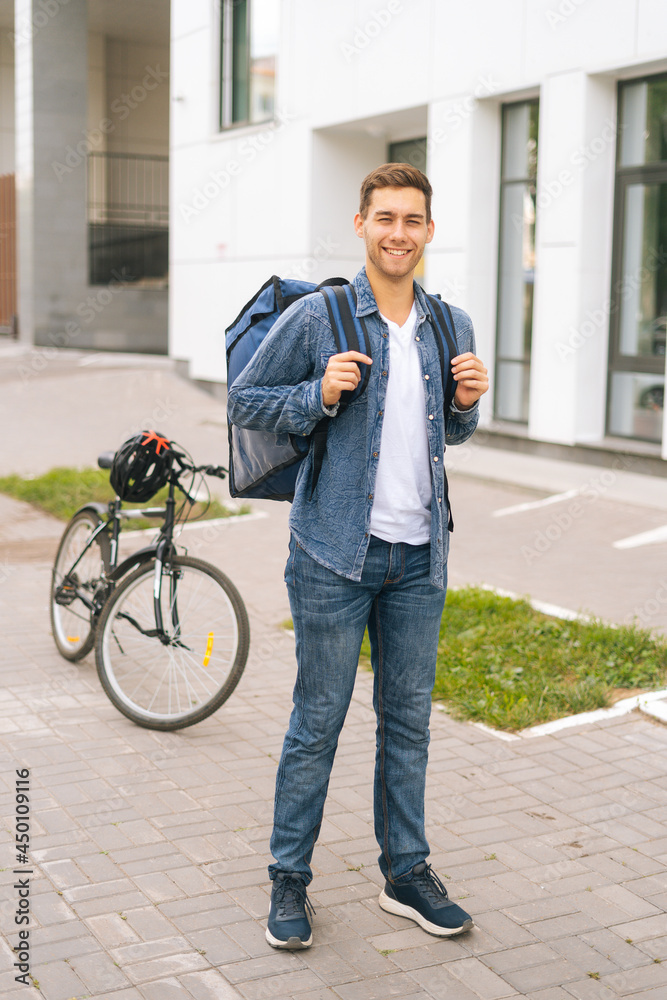 Fototapeta premium Full length vertical portrait of joyful handsome young delivery man with thermo backpack standing on city street, on background of bicycle and office building, looking at camera.