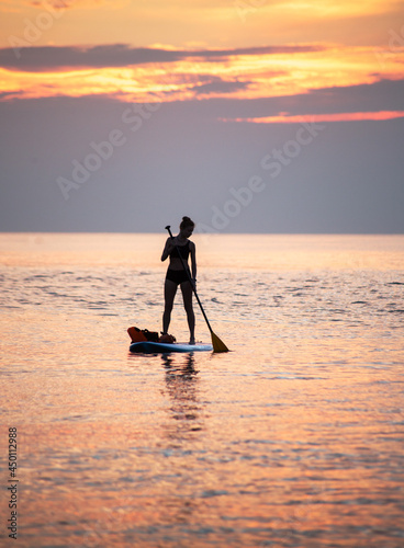 a woman is paddling on the SUP board at sea in the orange light of sunrise, a silhouette against a sunrise