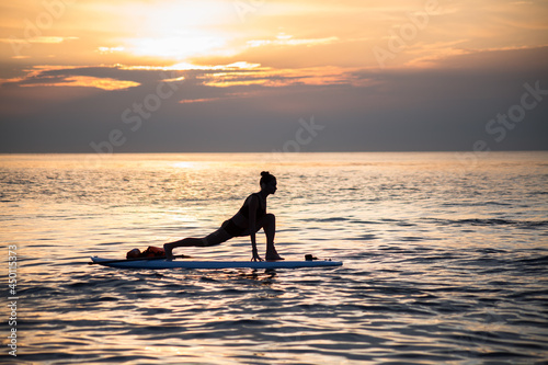 a female makes yoga asana on the SUP board at the orange light of sunrise