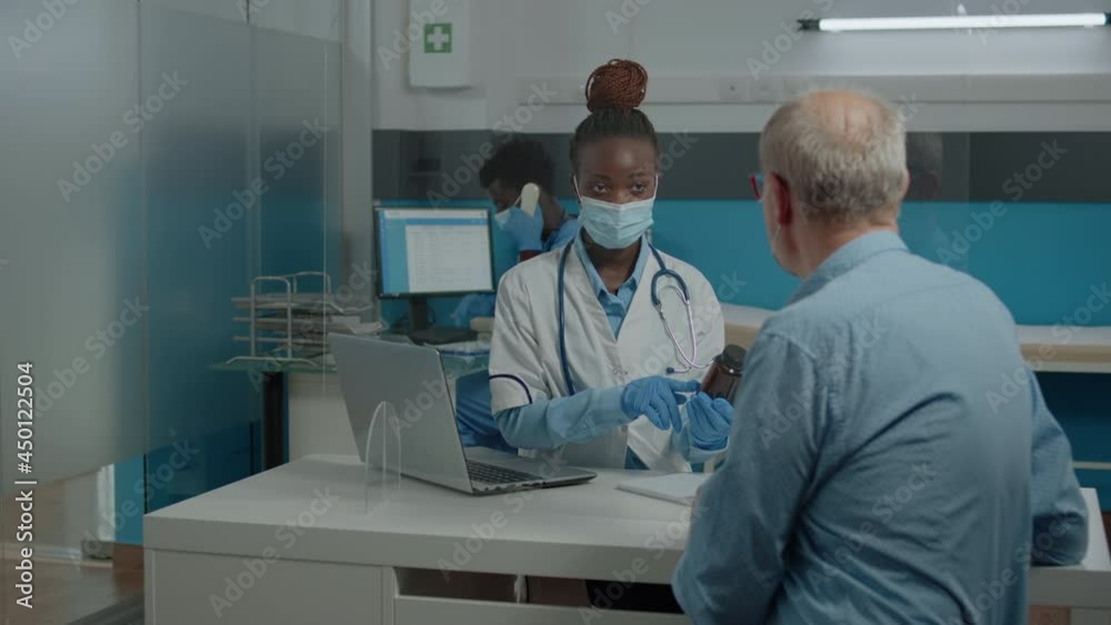 Black professional doctor showing bottle of pills to elder patient against sickness in medical office with glass protection. African american medic giving advice, and treatment to senior man Black professional doctor showing bottle of pills to elder patient against sickness in medical office with glass protection. African american medic giving advice, and treatment to senior man