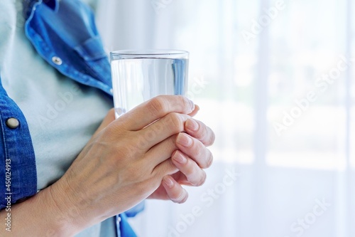 Close-up of middle-aged woman's hands holding glass of water, at home near window, copy space