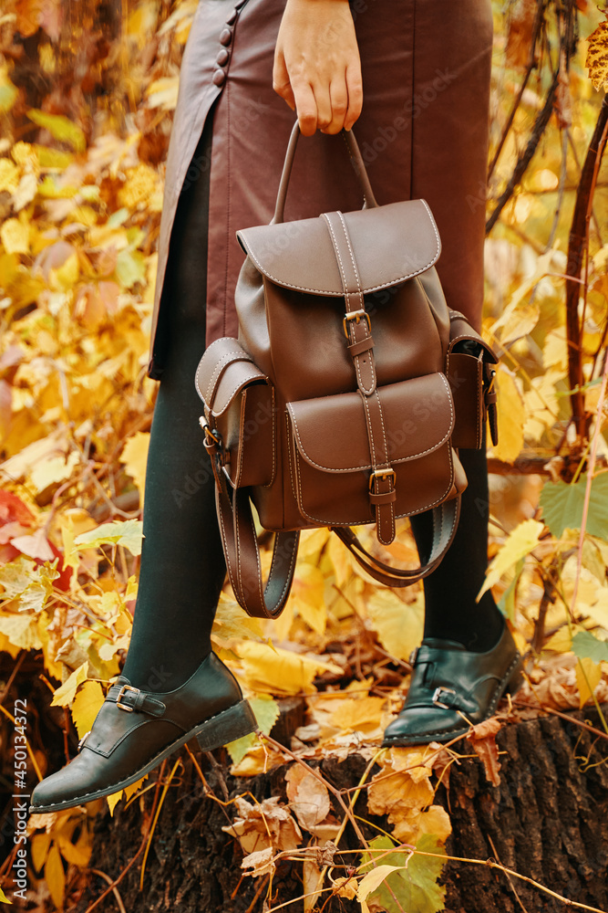 Fototapeta premium Pretty woman in skirt and with a backpack. Autumn walk in the Park. Fall foliage in the background. Nature of the forest. A woman stands on a stump.