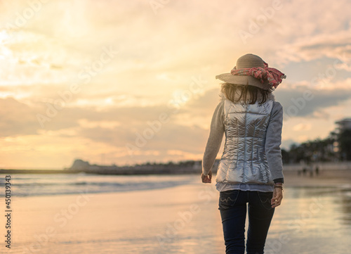 woman on her back with hat and scarf, walks along the beach at sunset.