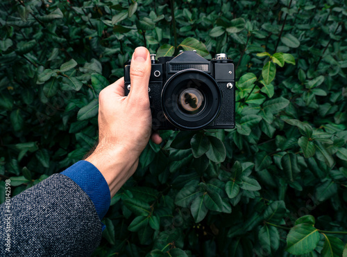 Male hand holding a vintage film camera in the air. Green nature in the background.