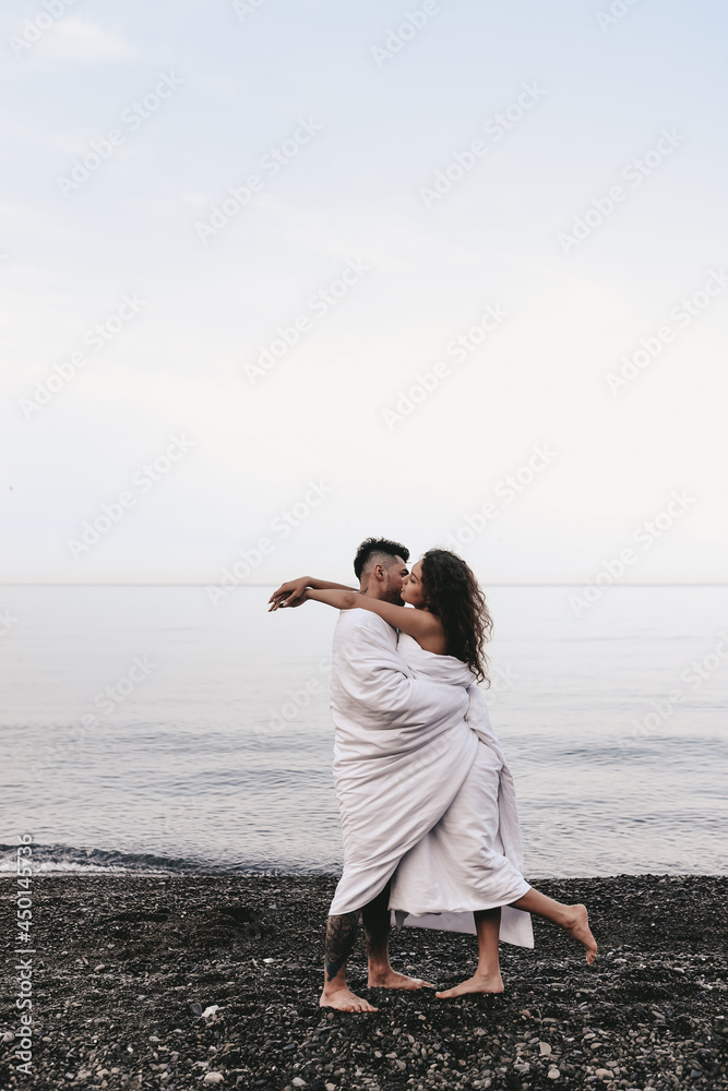 A couple in love are walking together hugging spend the night covered with a white blanket on the ocean beach on a romantic honeymoon in nature. Passionate man and woman of mixed diverse races A couple in love are walking together hugging spend the night covered with a white blanket on the ocean beach on a romantic honeymoon in nature. Passionate man and woman of mixed diverse races