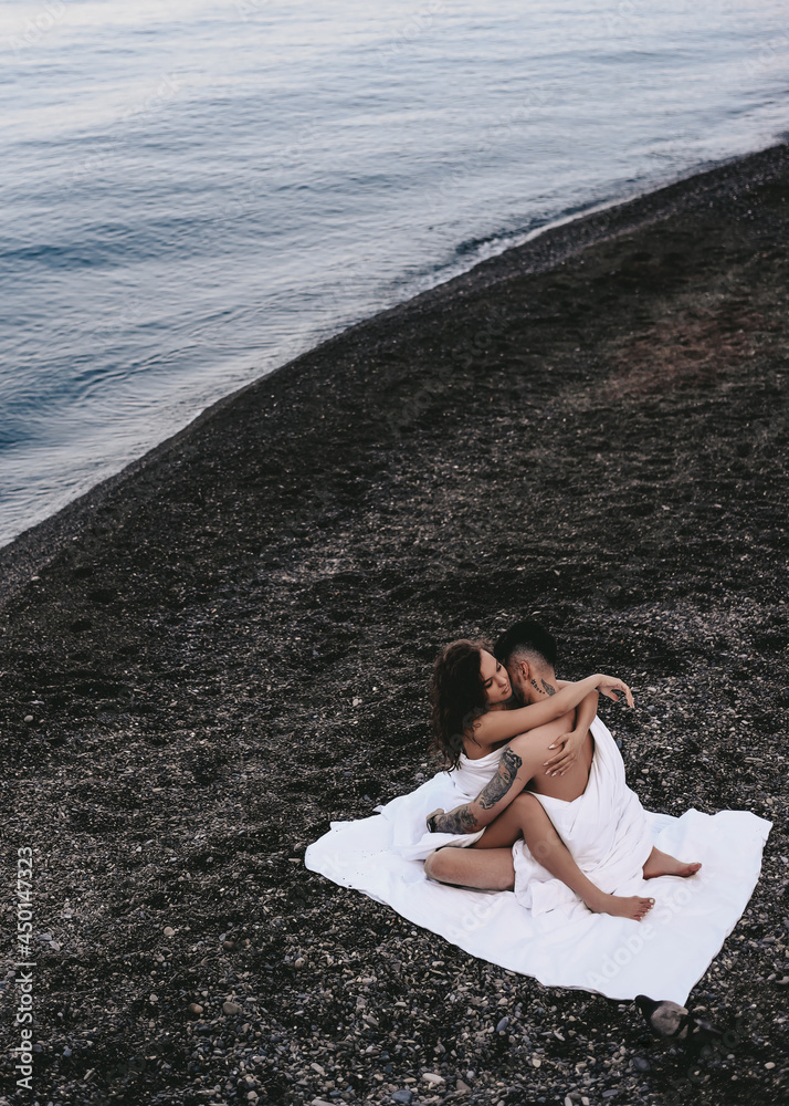 A couple in love are walking together hugging spend the night covered with a white blanket on the ocean beach on a romantic honeymoon in nature. Passionate man and woman of mixed diverse races A couple in love are walking together hugging spend the night covered with a white blanket on the ocean beach on a romantic honeymoon in nature. Passionate man and woman of mixed diverse races
