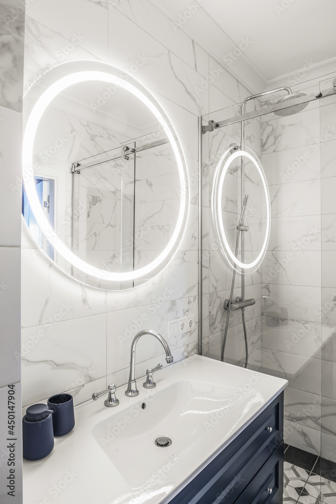 Modern bathroom interior with a dark blue wooden shelf, white sink and big round mirror with LED light. - obrazy, fototapety, plakaty 