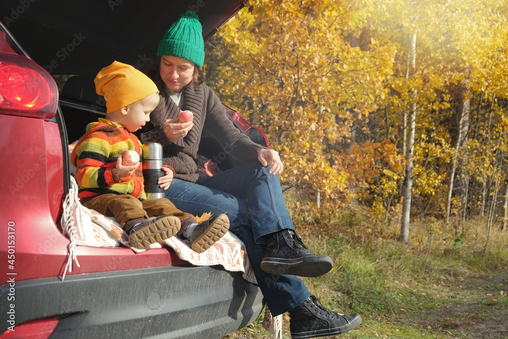 Family picnic outdoor, road trip in autumn season. Mother and her little child sitting inside car trunk near rural road in autumn forest and drinking tea from thermos.