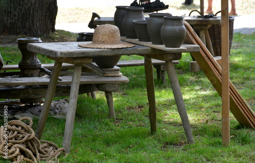 Close up on equipment of a medieval potter with a straw hat laying on a wooden table next to some pots, vases, and other utilitarian compartments made out of clay and colored with paint seen in Poland
