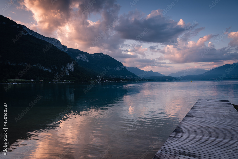 Natural landscape of Lake Annecy with colorful clouds at sunrise, Alps mountains in the background and a small wooden pier in the foreground, Annecy, France