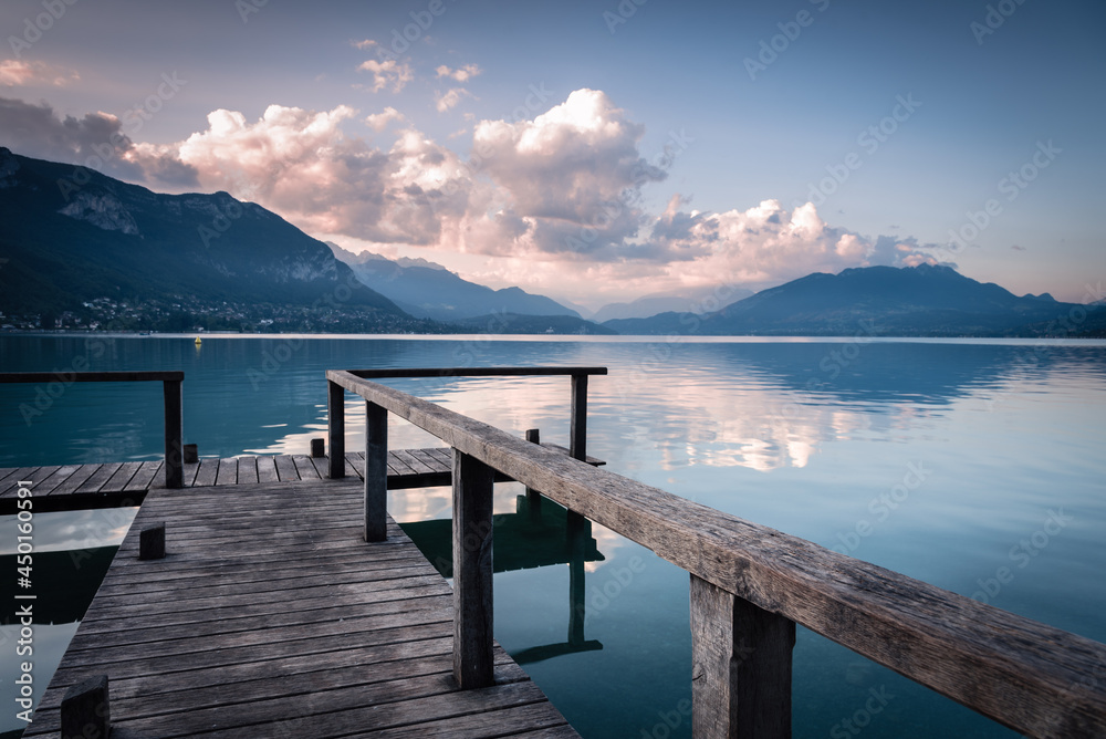 Natural landscape of Lake Annecy with colorful clouds at sunrise, Alps mountains in the background and a small wooden pier in the foreground, France