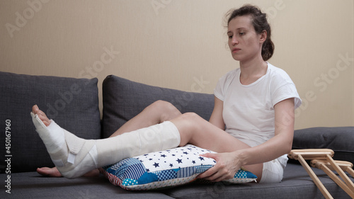 woman with a plaster cast sits on a sofa, a pillow is placed under a broken leg