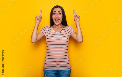 Cute excited smiling young brunette girl is pointing away for some advertisement while stands on yellow wall background