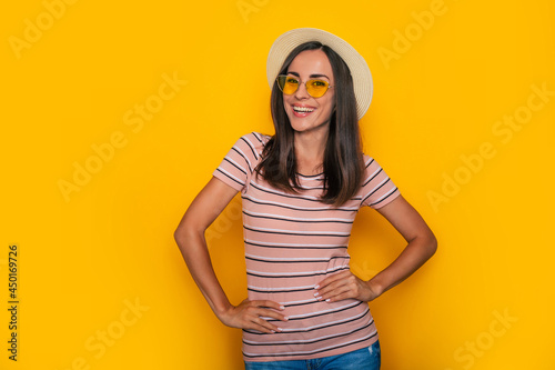 Happy excited beautiful tourist woman in hat and sunglasses is posing on yellow background and having fun