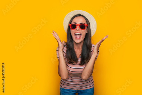 Happy excited beautiful tourist woman in hat and sunglasses is posing on yellow background and having fun