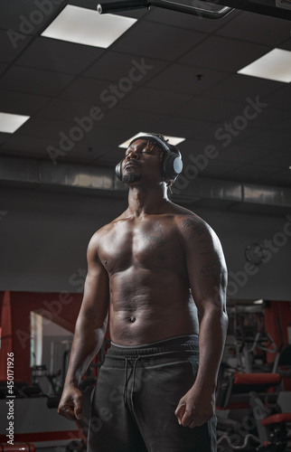 Athletic african american man in wireless headphones with closed eyes and naked torso standing in gym