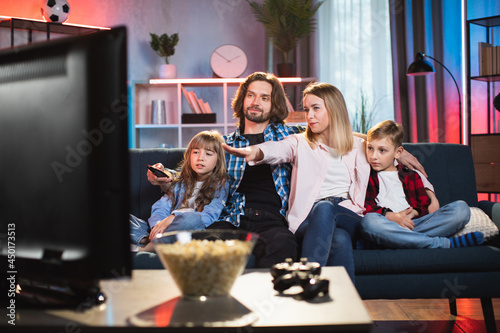 Caucasian parents with son and daughter sitting in embrace on sofa and watching movie during evening time. Young family in casual wear spending time together at home.